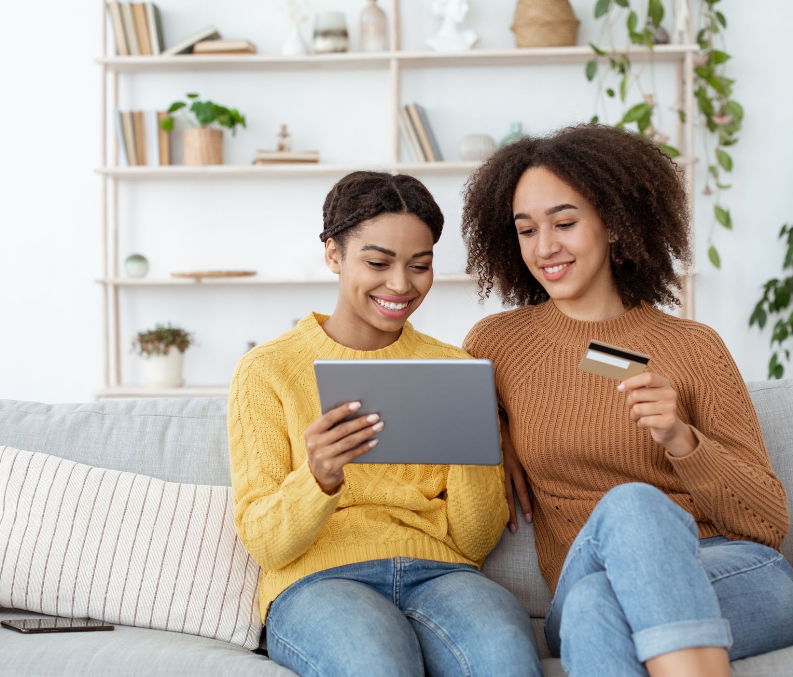 Two smiling women sitting on a couch holding a credit card and tablet