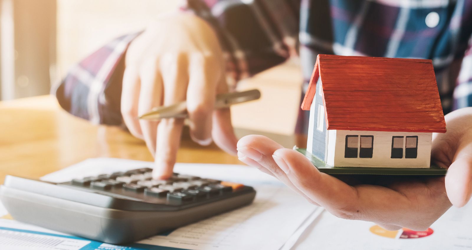 A man holding a small model home in one hand while typing into a calculator with the other hand